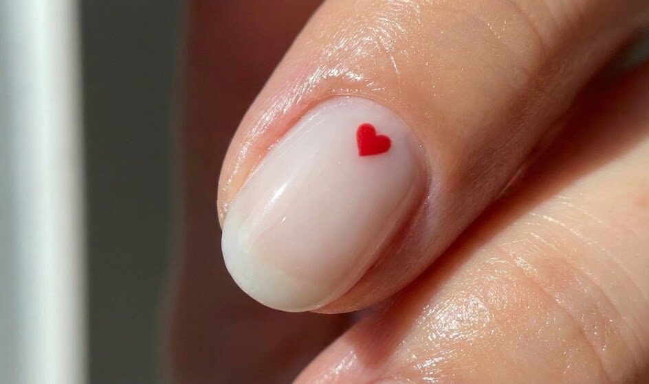 Makro zdjęcie paznokcia z minimalistycznym zdobieniem. Na mlecznej bazie widać jedno, malutkie czerwone serduszko