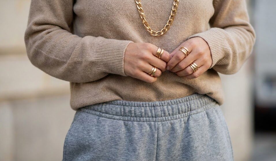 Zbliżenie na pas spodni dresowych i biżuterię. Kobieta ma włożony rąbek swetra w spodnie, widać złoty łańcuch na szyi i kilka pierścionków. Zbliżenie na pas spodni dresowych i biżuterię. Kobieta ma włożony rąbek swetra w spodnie, widać złoty łańcuch na szyi i kilka pierścionków.