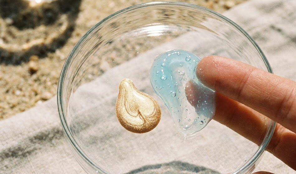 Zbliżenie na teksturę kosmetyków na szkle lub dłoni. Widać kroplę złotego, płynnego rozświetlacza oraz lekką, żelową konsystencję kremu